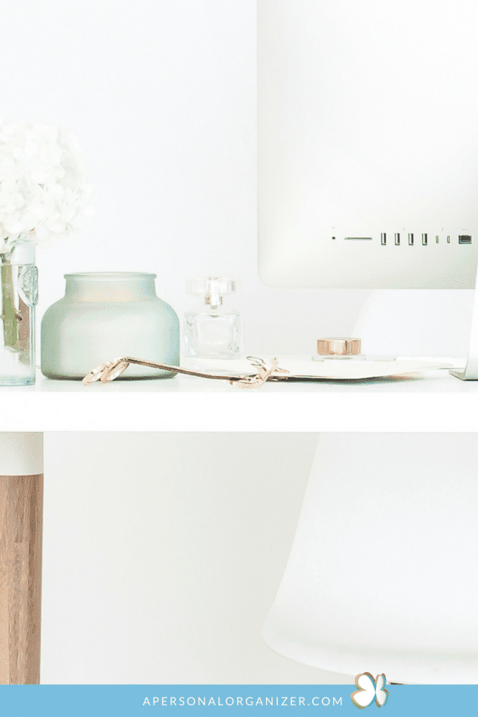 A minimalist white desk setup featuring an iMac, a small vase with white flowers, a green candle holder, a perfume bottle, and a pair of glasses. The desk has a wooden leg and a white chair. The scene is bright and clean, with neatly organizing the cords beneath ensuring no clutter, and website text at the bottom.