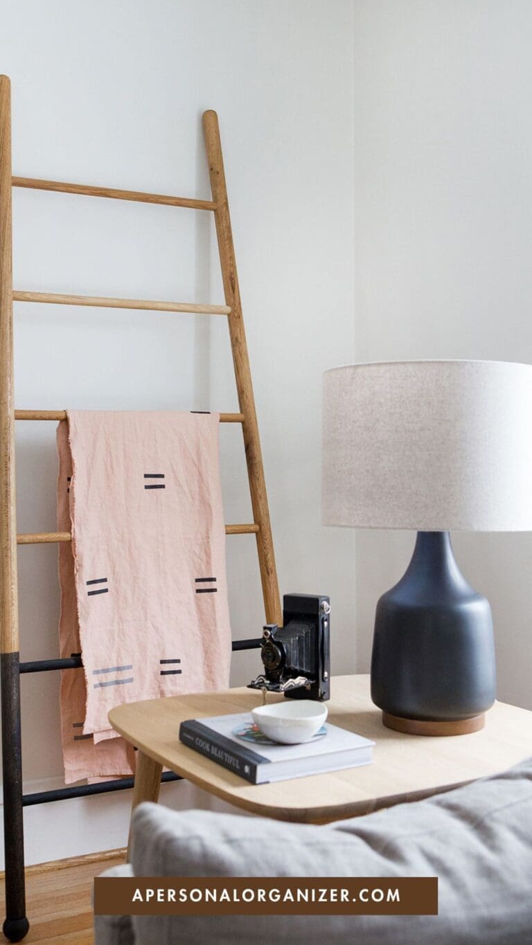 A minimal, modern room features a wooden ladder with a pink cloth, a small table with a gray lamp, vintage camera, book, and bowl—perfect for stylish organization. The website "apersonalorganizer.com" is displayed at the bottom.