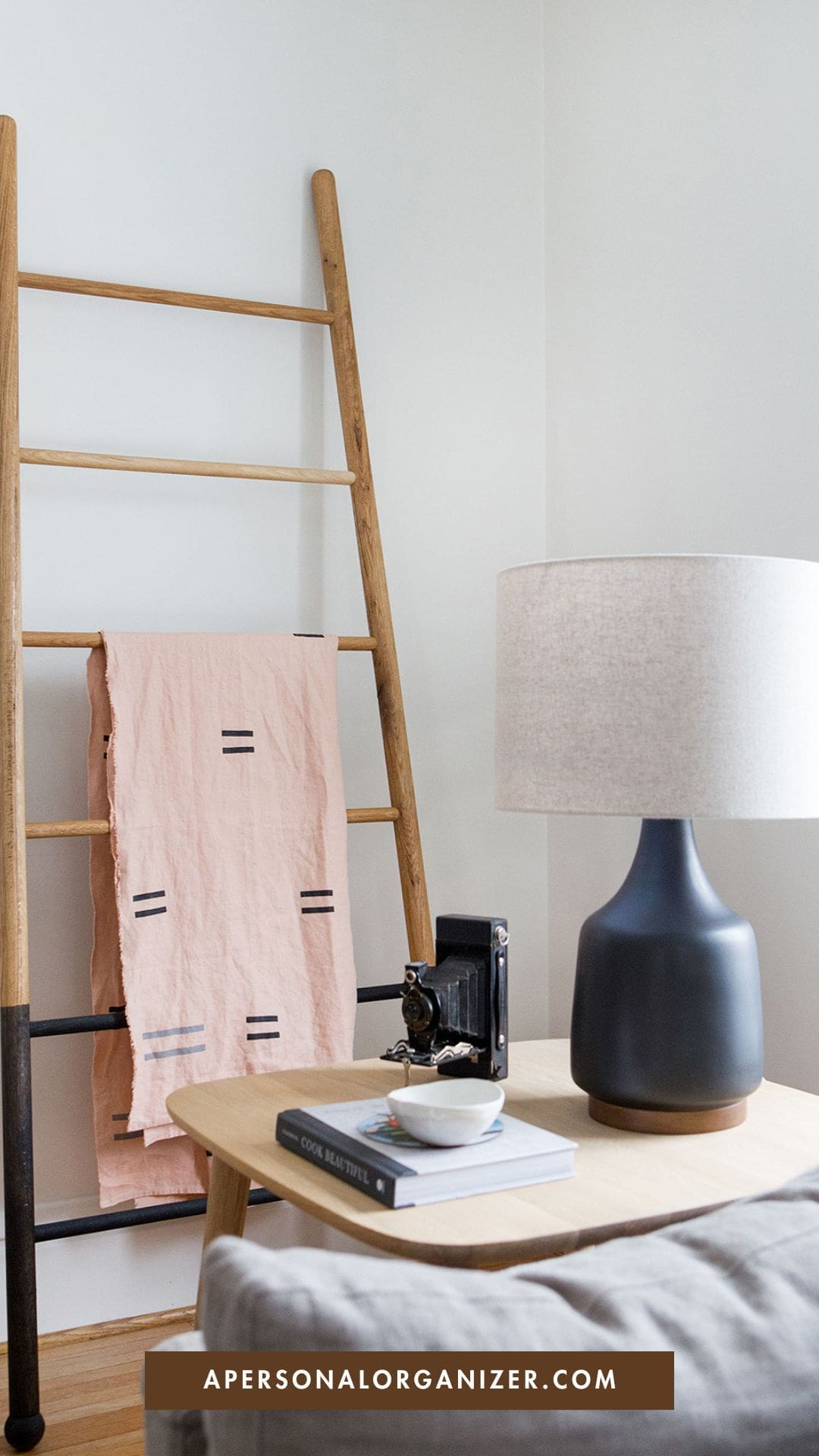 A minimal, modern room features a wooden ladder with a pink cloth, a small table with a gray lamp, vintage camera, book, and bowl—perfect for stylish organization. The website "apersonalorganizer.com" is displayed at the bottom.