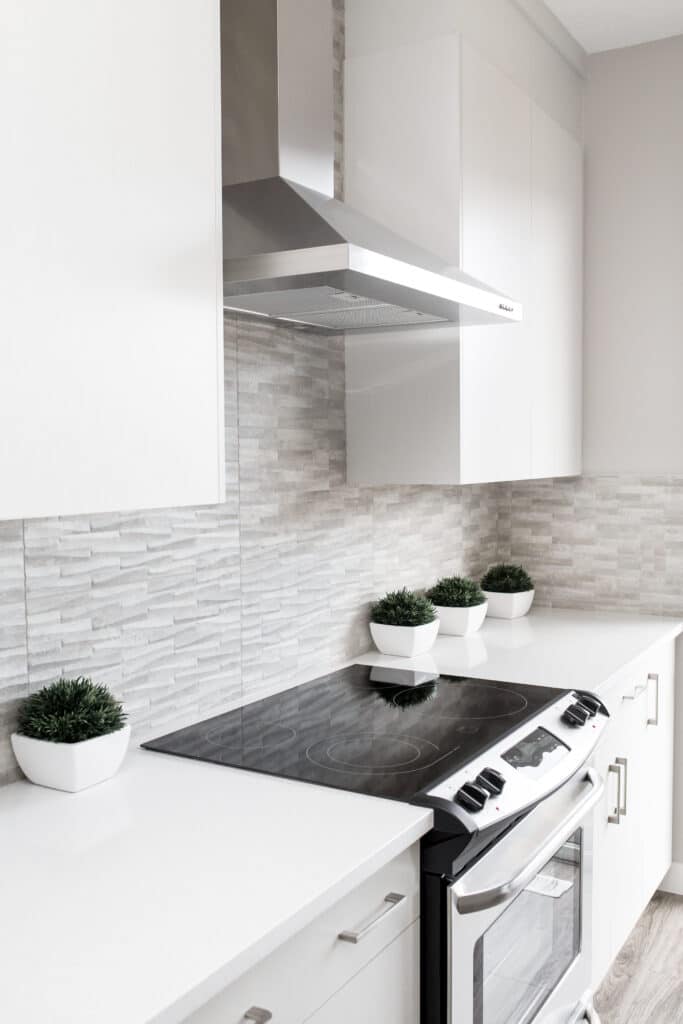 A modern kitchen with white cabinetry, stainless steel stove and hood, and a sleek black cooktop. The backsplash features light grey textured tiles, making the space easy to incorporate into your cleaning routine. Four small potted plants in white square pots are neatly lined up on the counter.