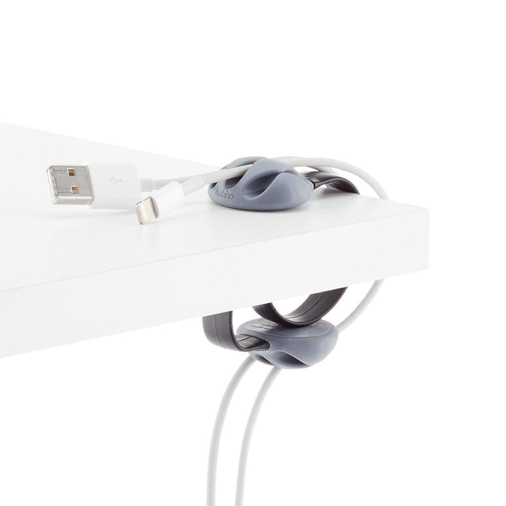 A white desk features two grey cable organizers, perfect for organizing the cords, holding white charging cables neatly in place at the edge. One of the cables has a USB plug end resting on the desk surface. The desk and organizers are set against a plain white background.