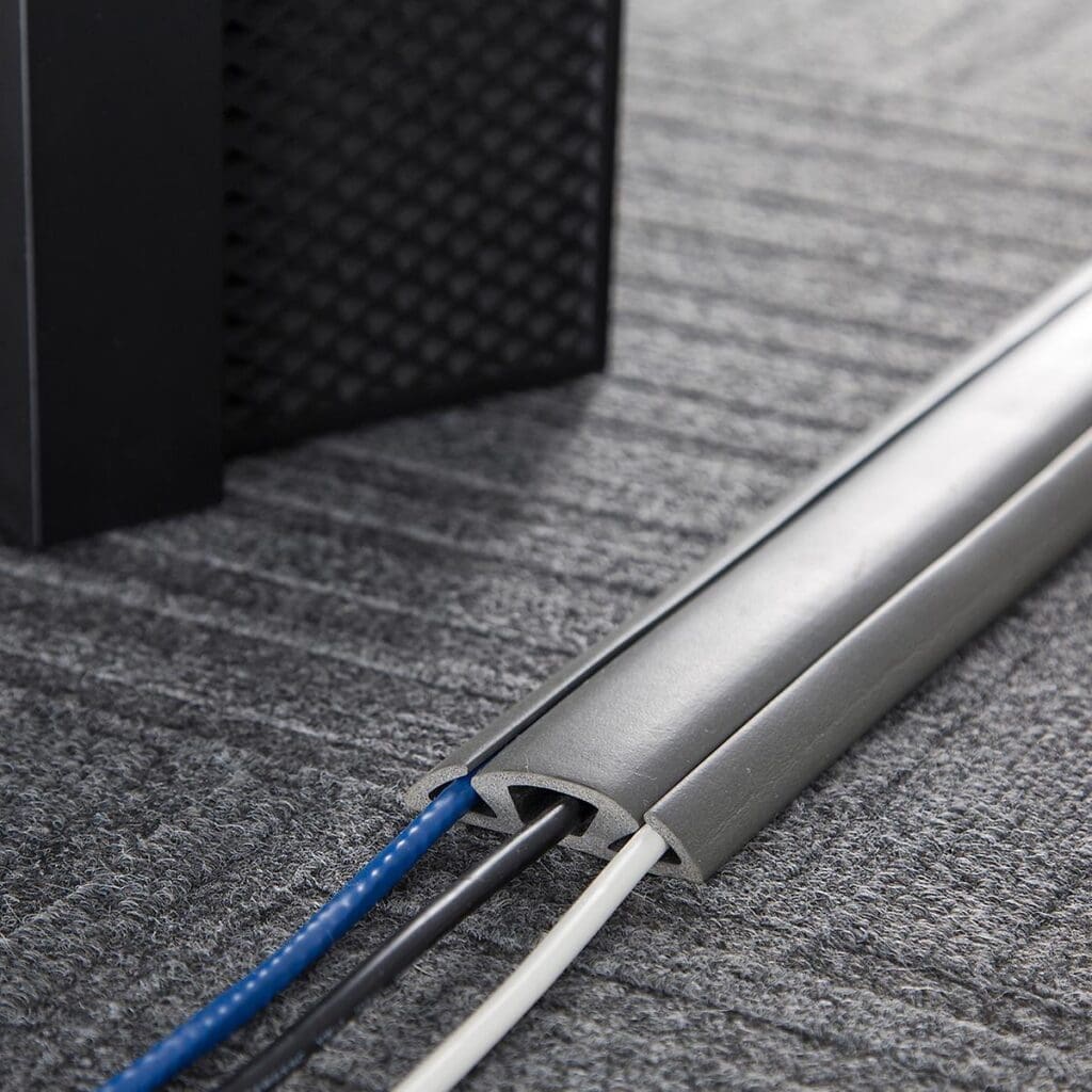 A few cables, including blue and white ones, are neatly organized and covered by a grey cable management sleeve on a grey carpeted floor. The setup emphasizes organizing the cords efficiently. Part of a black piece of furniture is visible in the background.