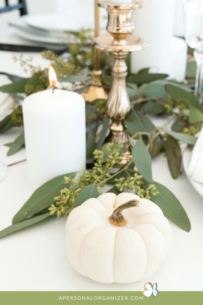 A white pumpkin is placed on a decorated table with greenery, a lit white pillar candle, and a silver candlestick. The scene, devoid of clutter, suggests a festive, autumnal or Thanksgiving setting. A website URL "apersonalorganizer.com" is visible at the bottom of the image.