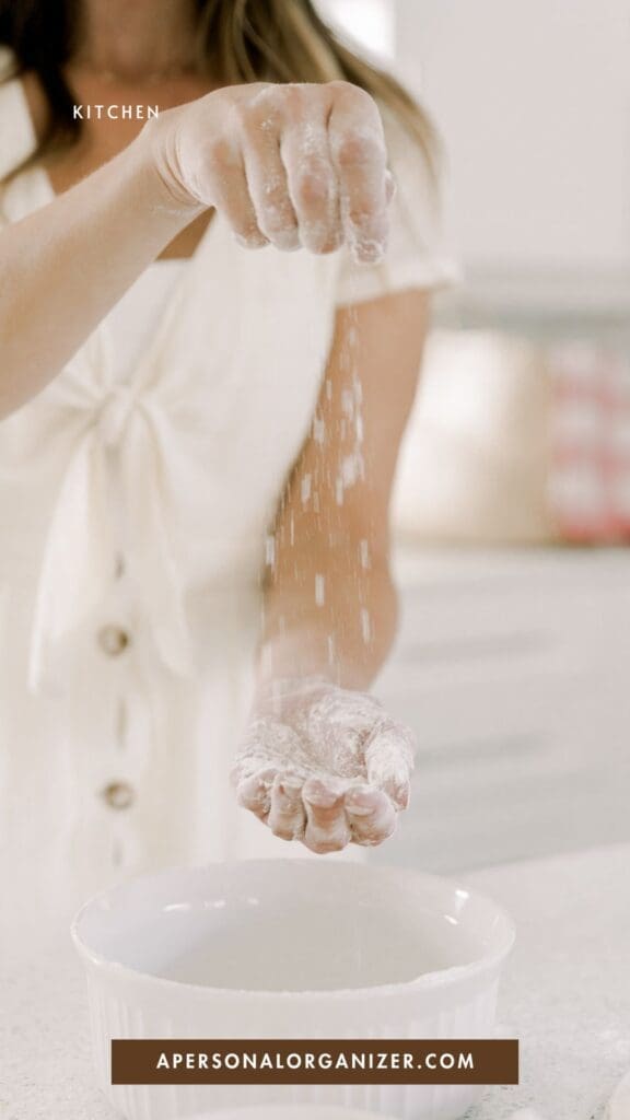 A woman in a white dress sprinkles flour into a white bowl at her baking station in a bright kitchen. The text on the image reads "KITCHEN" and "APERSONALORGANIZER.COM".