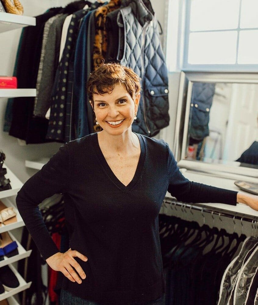 A woman with short brown hair, wearing a black long-sleeve top, stands smiling in a neatly organized closet—an inspiring setting for a curso personal organizer—with natural light streaming through a window onto tidy shelves and hanging clothes.