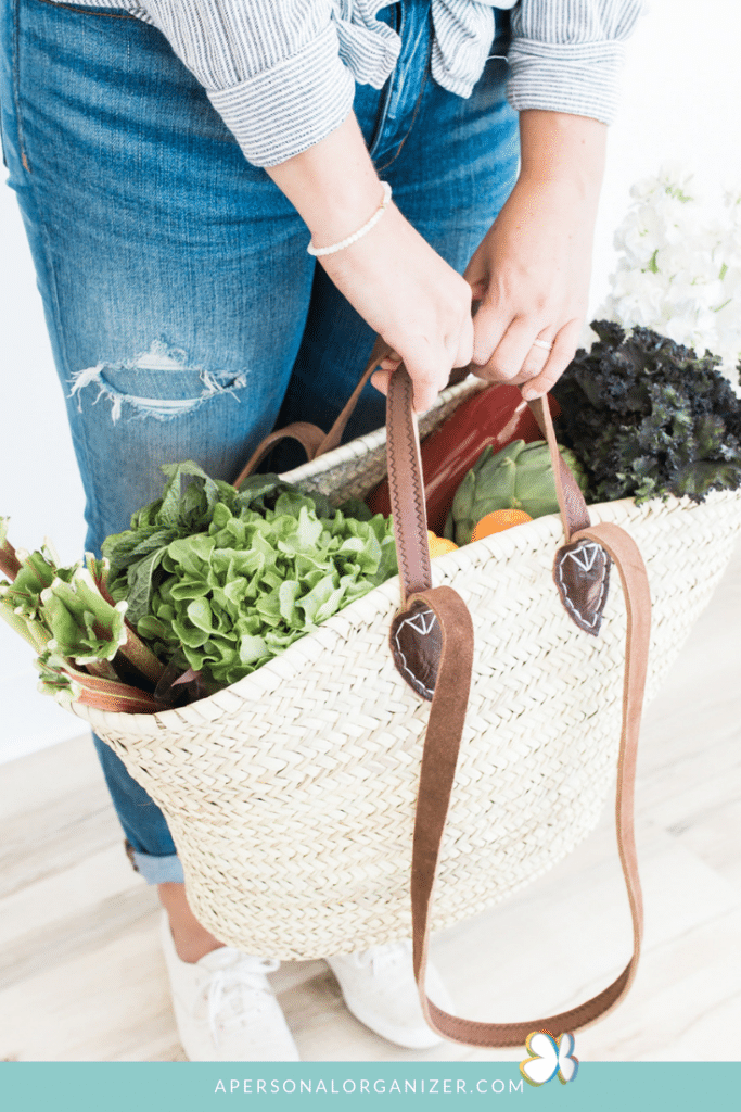 A person in a blue and white striped shirt and ripped jeans is holding a woven basket filled with fresh greens and vegetables, perfect for meal planning. The basket has brown leather handles. A logo and URL "apersonalorganizer.com" are visible at the bottom of the image.
