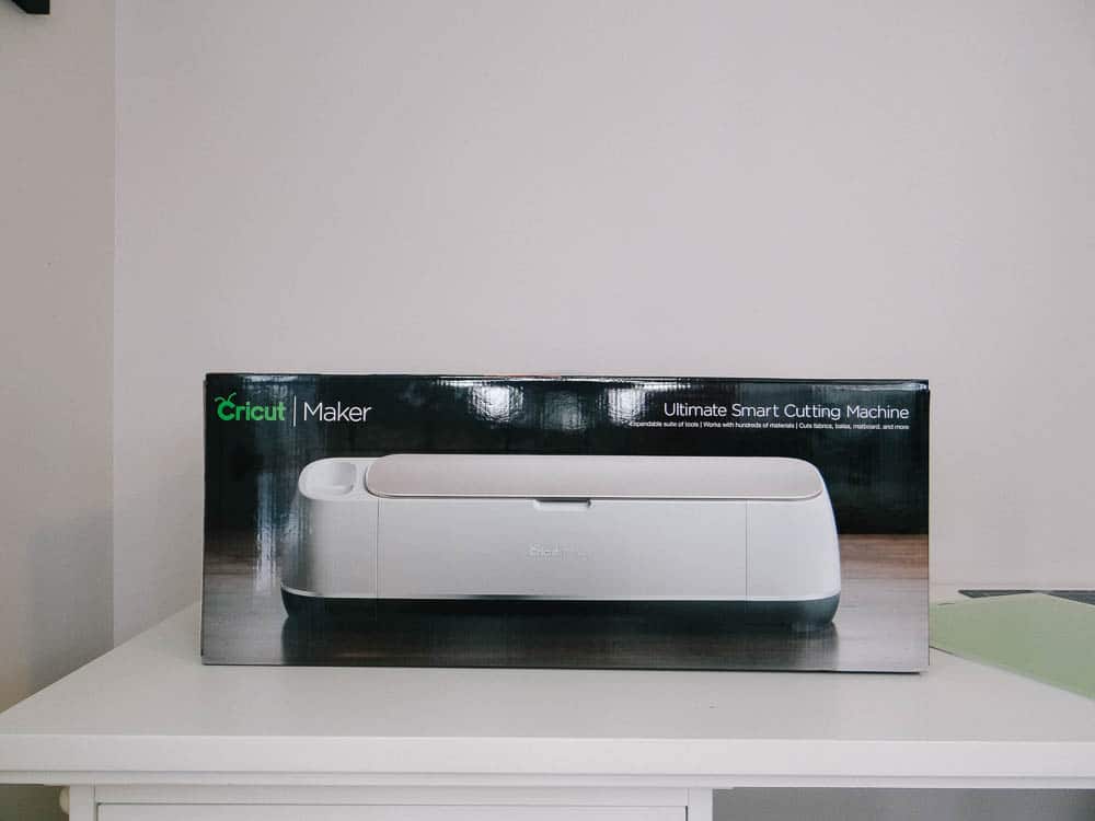 The image showcases a boxed Cricut Maker, labeled as the "Ultimate Smart Cutting Machine." The box features a picture of the sleek white Cricut Maker, designed for various crafting and cutting tasks. It rests on a white surface in a well-lit room.