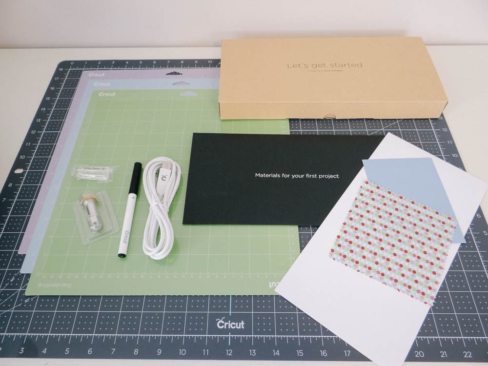 A Cricut Maker cutting mat with various items laid out on it, including a box labeled "Let's get started," a black pen, a white cord, a small blade in a plastic case, two cutting mats, a black envelope with "Materials for your first project," and patterned paper.