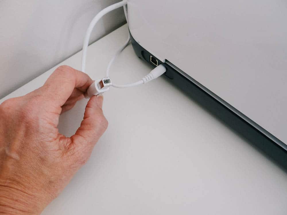 A hand is seen plugging a USB cable into the port of a device, possibly a Cricut Maker or a similar electronic device, which is placed on a white table. The focus is on the connectors and the action of connecting the cable.