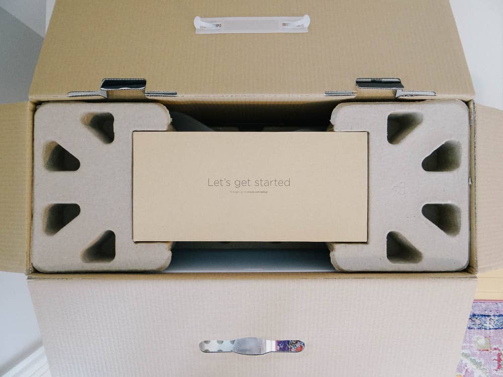 An open cardboard box revealing its contents: a beige rectangular Cricut Maker box with the text "Let's get started" on top, nestled securely between two triangular cardboard spacers. The flaps of the main box are folded open.
