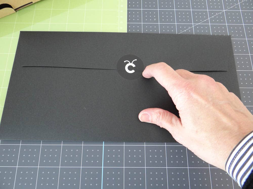 A hand in a striped cuff shirt reaches to open a black envelope sealed with a circular sticker featuring a white logo, set against a background of crafting and cutting mats alongside the Cricut Maker.