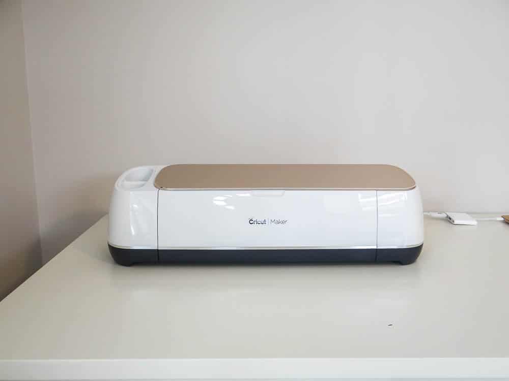A Cricut Maker machine sits on a white table against a light-colored wall. The device has a sleek, rectangular design with a white base and a tan top cover. The Cricut Maker logo is visible on the front, and a power cable is plugged into the right side.