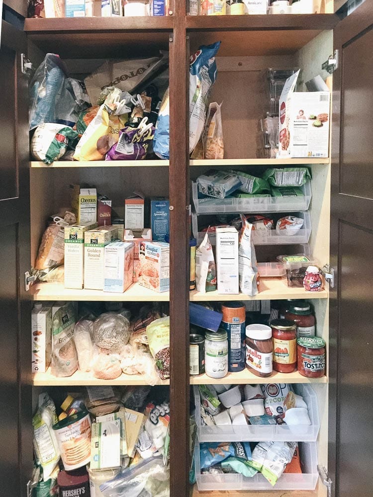 A cluttered pantry with its doors open, showing an assortment of food items and snacks. Shelves are filled with boxes, jars, bags, and canned goods, including cereal, pasta, sauces, and baking ingredients. Among the chaos sits a Cricut Maker box wedged between stacks of disorganized items.