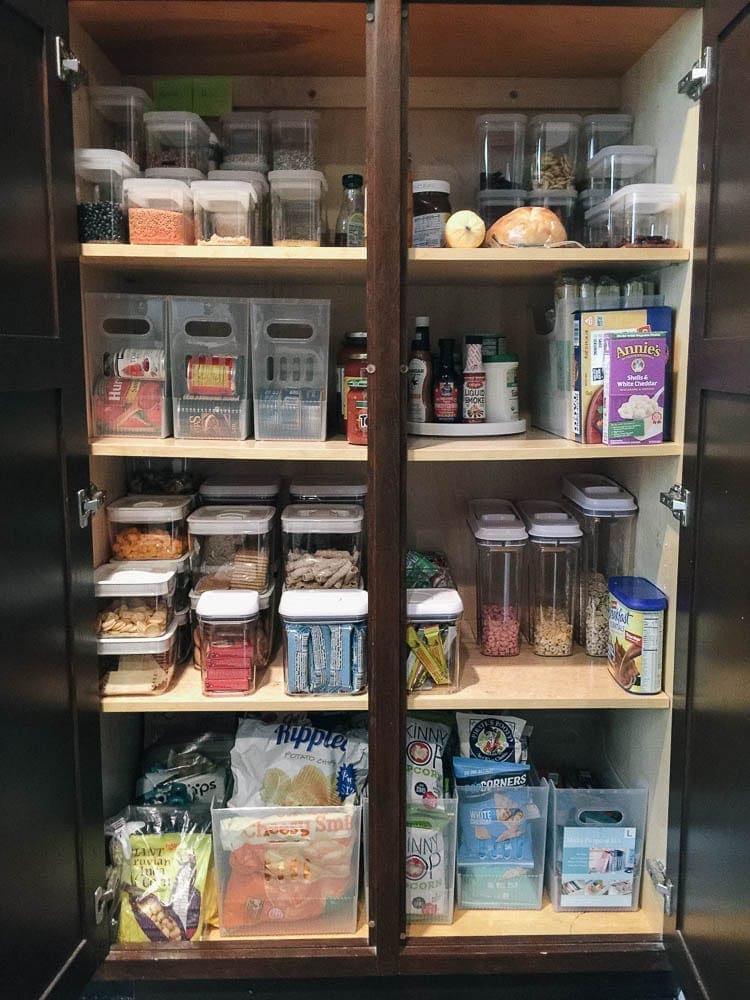 A neatly organized pantry with labeled clear containers on shelves, crafted using a Cricut Maker, filled with various food items, including grains, snacks, sauces, and seasonings. Boxes, jars, and packages are also visible, contributing to the orderly arrangement.