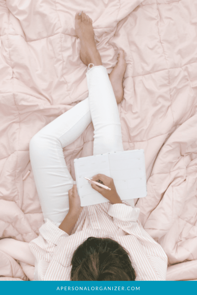 A person sits on a pink quilted blanket, wearing white pants and a striped top, writing in a notebook with a pen. Their feet are bare, and they are viewed from above. A blue banner at the bottom reads "APERSONALORGANIZER.COM"—your go-to resource to declutter your personal life.