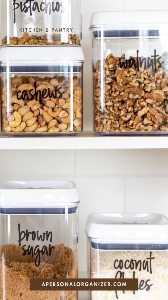 Four clear food storage containers on white shelves, labeled with Cricut vinyl labels and filled with pistachios, walnuts, cashews, brown sugar, and coconut flakes. A website label at the bottom reads "apersonalorganizer.com.
