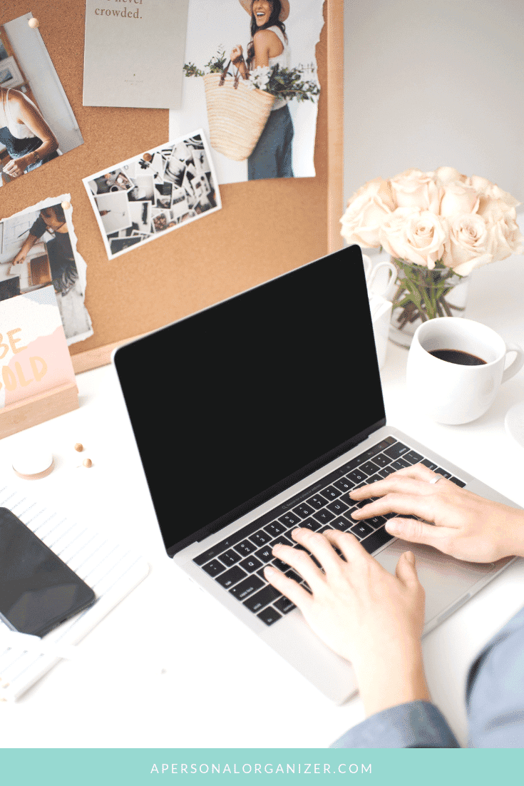 A person typing on a laptop at a bright, decorated workspace. The desk includes a cup of coffee, a notebook, and a smartphone, hinting they might be about to start a blog. A corkboard displays photos, notes, and a small vase of flowers in the background. The bottom edge of the image reads "apersonalorganizer.com".