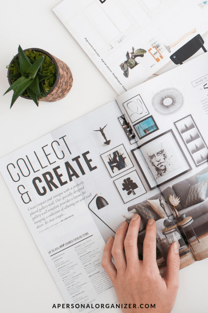 A hand holding a magazine open to a page titled "COLLECT & CREATE." The page displays a stylishly decorated room with various wall art and decor pieces, offering inspiration akin to blog content strategy. A small plant and part of another magazine are visible on the white surface next to the open magazine.