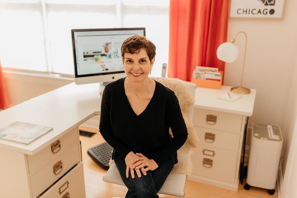 A person with short brown hair sits in a home office, smiling at the camera. They wear a black sweater and jeans. Behind them, an organized desk reflects their knack for office organizing, featuring a computer, a magazine, and essential supplies. Bright red curtains and white walls are also visible.