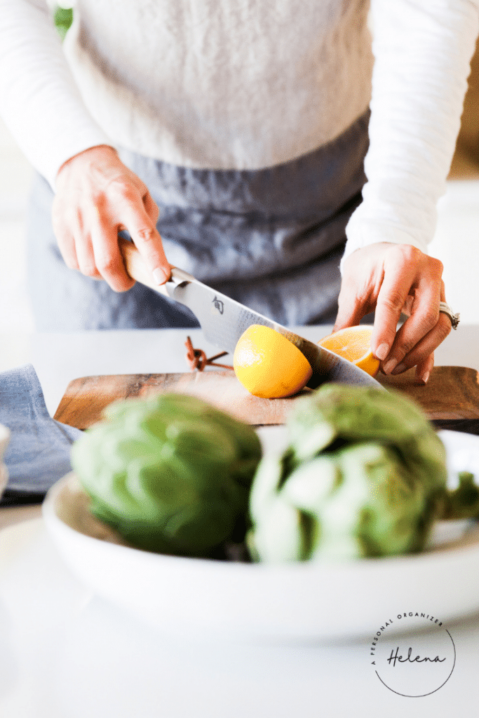A person, embodying everyday organizing solutions, slices a lemon on a wooden cutting board. Artichokes sit elegantly on a white plate in the foreground. Dressed in a light-colored top and gray apron, they work efficiently under the "Helena" logo prominently displayed in the corner.