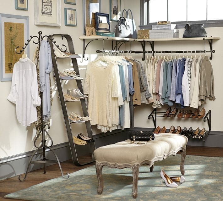 A neatly organized walk-in closet featuring hanging clothes in neutral and pastel colors, shelves with shoes and handbags, a cushioned bench in the center, framed art on the walls, and smart closet organizing ideas throughout.