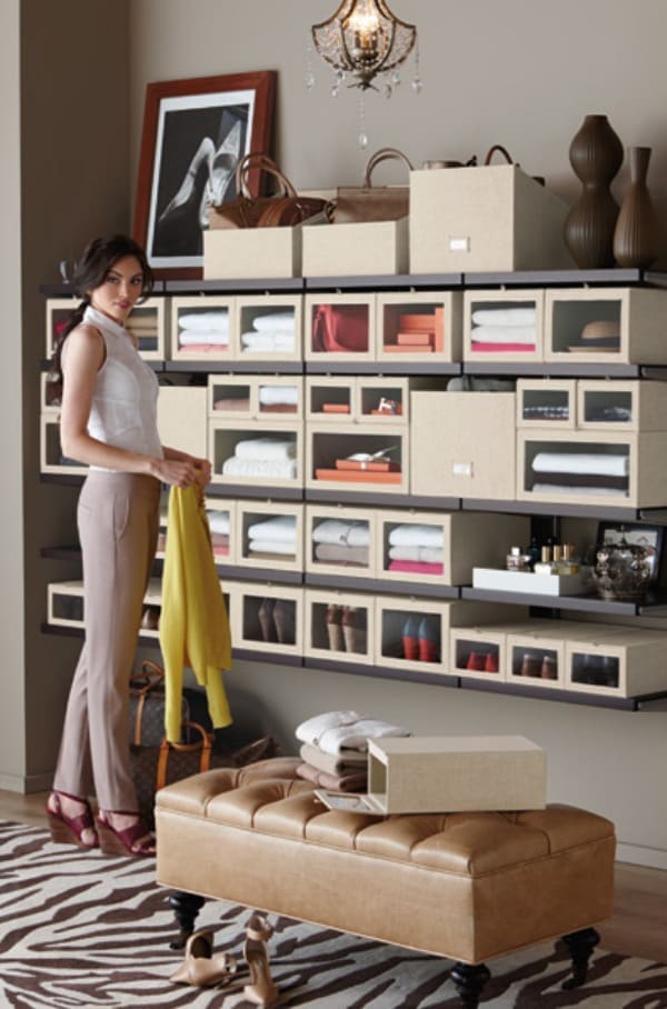 A woman stands near wall-mounted shelves organized with labeled boxes holding clothes, shoes, and accessories. Showcasing clever closet organizing ideas, the stylish room features a bench, framed photo, vases, and a chandelier.