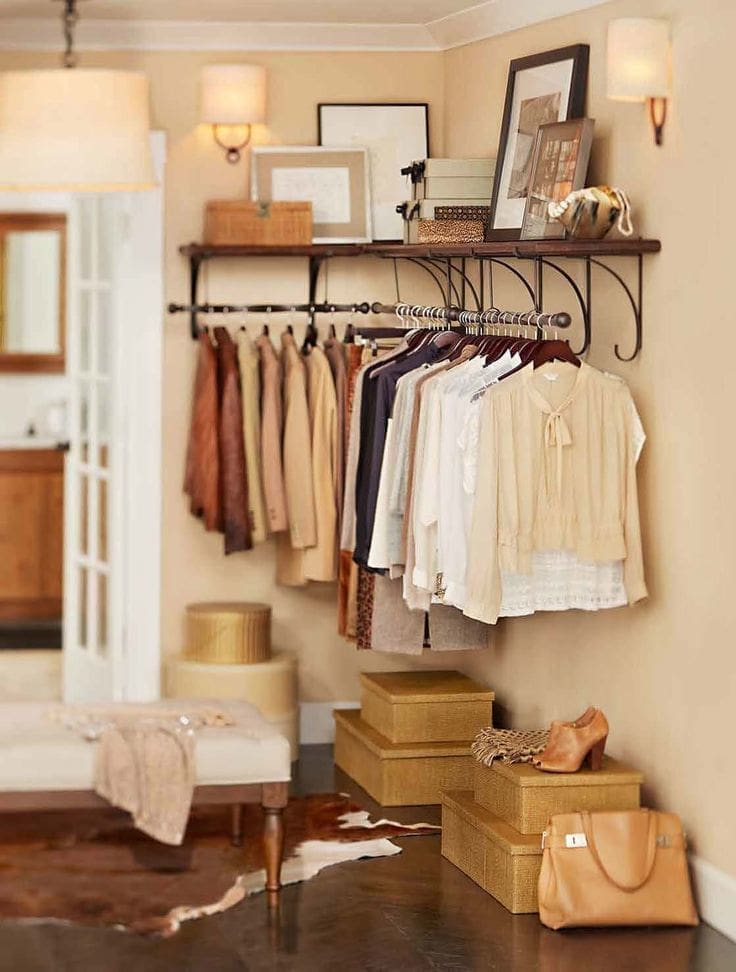 Cozy bedroom corner with beige and white clothes elegantly displayed on a rack, showcasing practical closet organizing ideas. Various accessories rest on the shelf above, while boxed storage and a handbag sit neatly on the floor. A small bench and soft lighting complete the warm, inviting atmosphere.