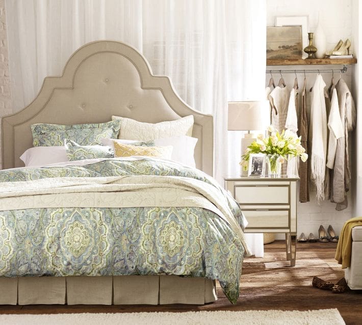 A cozy, elegant bedroom with a tufted beige headboard, patterned bedding in soft blue and cream, white sheer curtains, a mirrored nightstand with a lamp and flowers, and neutral clothes neatly displayed for closet organizing ideas.