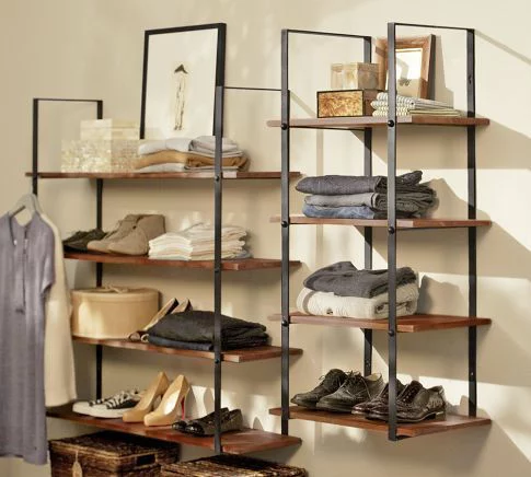 A modern shelving unit with a dark metal frame and wooden shelves showcases closet organizing ideas by holding folded clothes, shoes, baskets, and decorative items. A dress hangs elegantly on the side. The neutral color scheme enhances its organized and minimalist style.