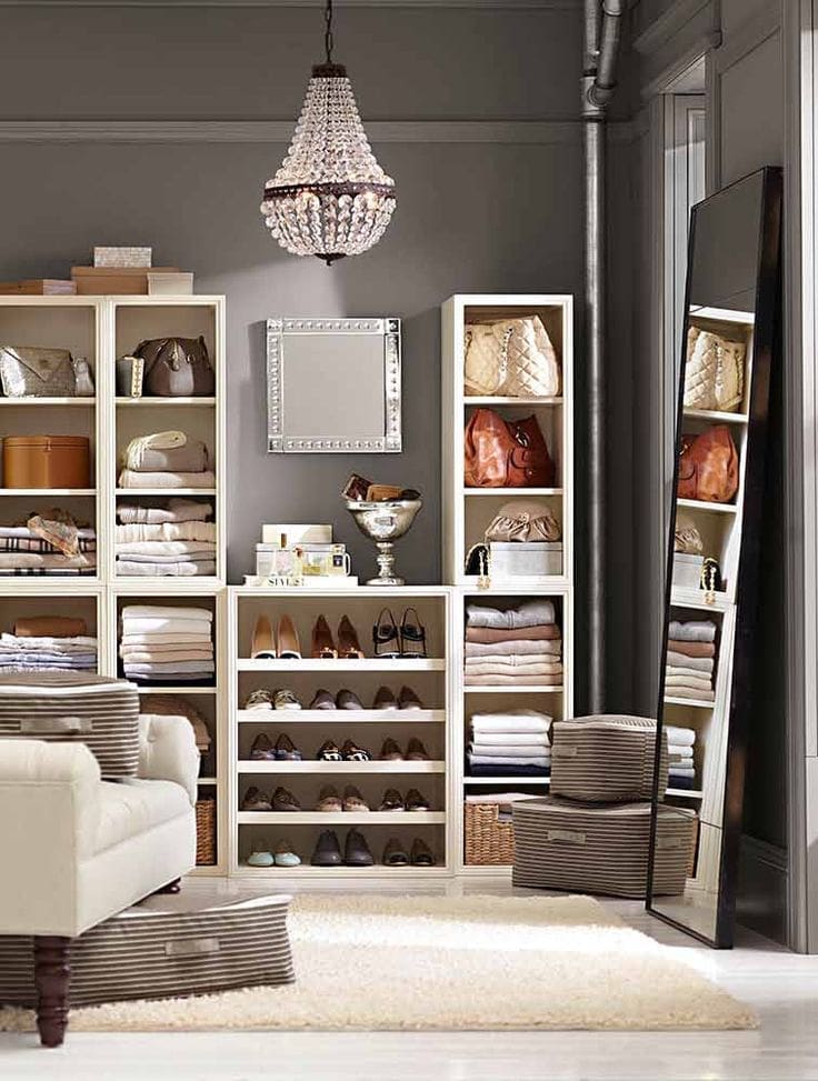 A neatly organized walk-in closet with shelves holding folded clothes, handbags, and shoes. Showcasing smart closet organizing ideas, a chandelier hangs above, a large mirror leans against the wall, and an armchair sits on a plush rug.