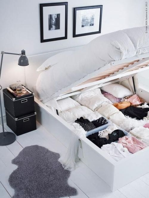A white bed with the mattress lifted reveals organized storage underneath—perfect for folded clothes and pillows. The bright room features minimal decor, a lamp, framed art, and black storage boxes for clever closet organizing ideas.