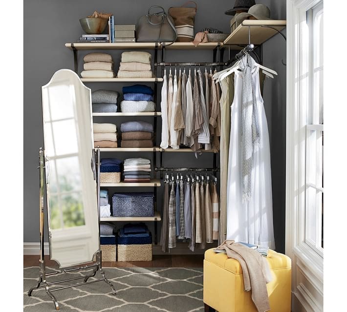 A neatly organized closet showcases clever closet organizing ideas with folded clothes on shelves, hanging shirts, and a long dress. A freestanding mirror stands nearby. A yellow ottoman sits on a patterned rug in front of a large window, letting in natural light.