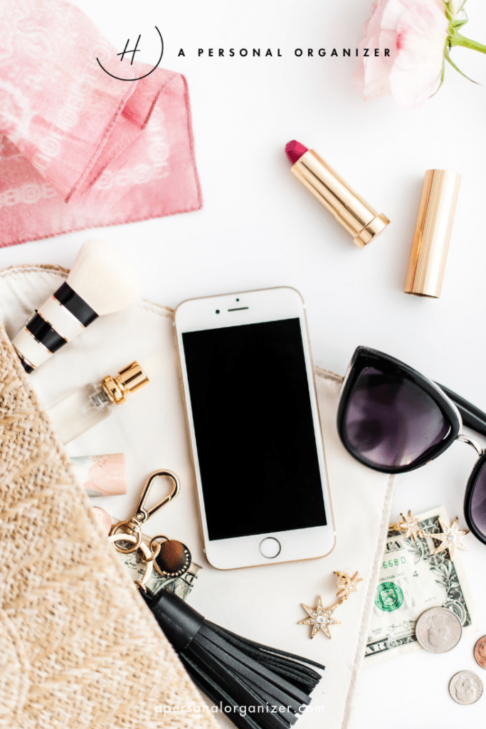 A flat lay image showcases a smartphone, sunglasses, and lipstick beside a makeup brush and star-shaped earrings on a white surface. Amid the organized commitments of fashion are a pink scarf, straw bag, gold keychain, dollar bills, and a chic tassel accessory.