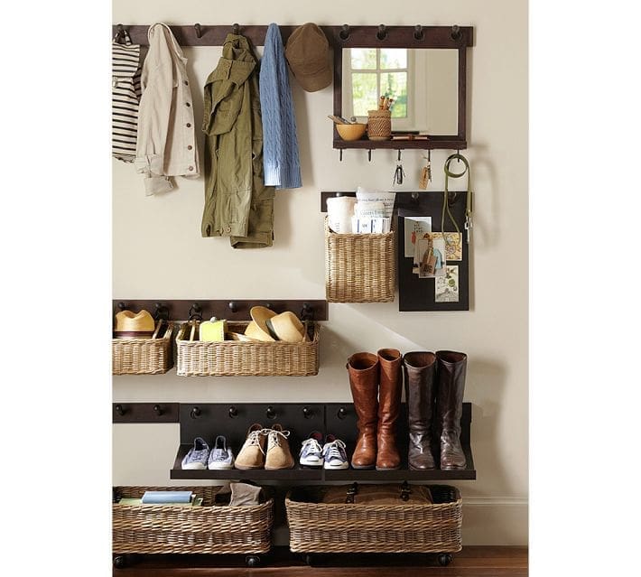 Entrance solution for organizing a narrow entry: wall hooks hold jackets, scarves, and a bag; a mirror sits above a basket for mail and keys; shelves below feature wicker baskets with shoes, hats, and boots neatly organized.