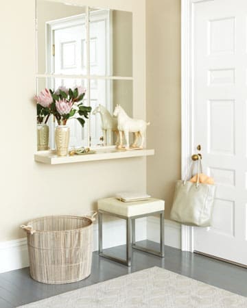 A tidy entrance designed for organizing a narrow entry, featuring a white door, beige walls, a mirror, floating shelf with vases and flowers, horse figurines, woven basket, small stool, and a light bag hanging on a hook.