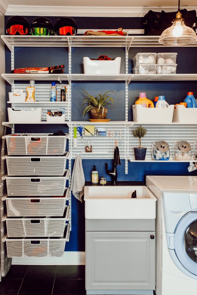 A well-organized laundry room features a white shelving unit neatly holding storage bins, detergent bottles, and cleaning supplies, making organizing the laundry room effortless. A small sink with a cabinet sits beside the washing machine. Dark blue walls and a plant add greenery.