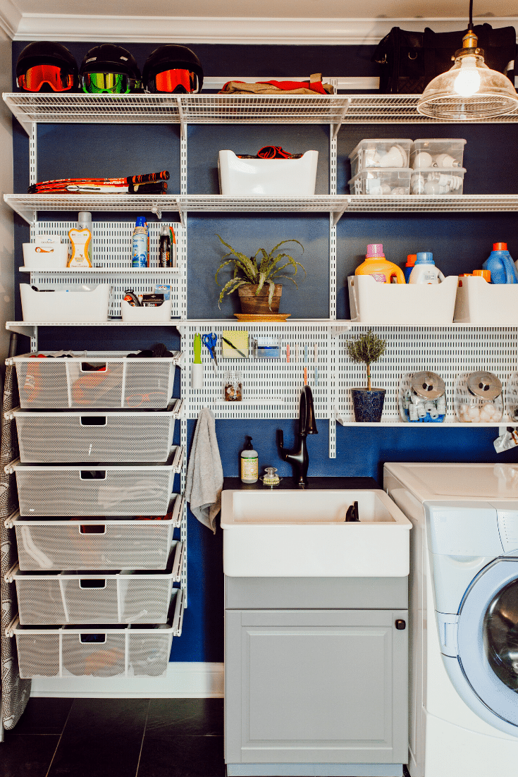 A well-organized laundry room features a white shelving unit neatly holding storage bins, detergent bottles, and cleaning supplies, making organizing the laundry room effortless. A small sink with a cabinet sits beside the washing machine. Dark blue walls and a plant add greenery.