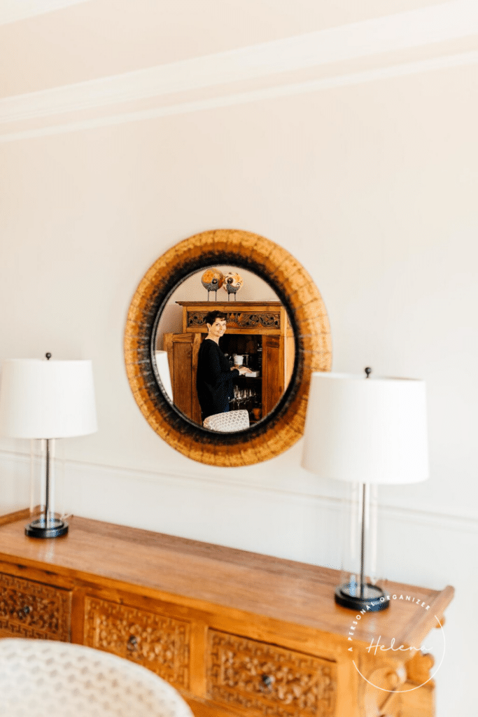 A round mirror reflects a person standing in front of a wooden cabinet, organizing the dining room. The mirror is mounted above a carved wooden console table with two lamps on either side. The room is softly lit, with neutral wall colors.
