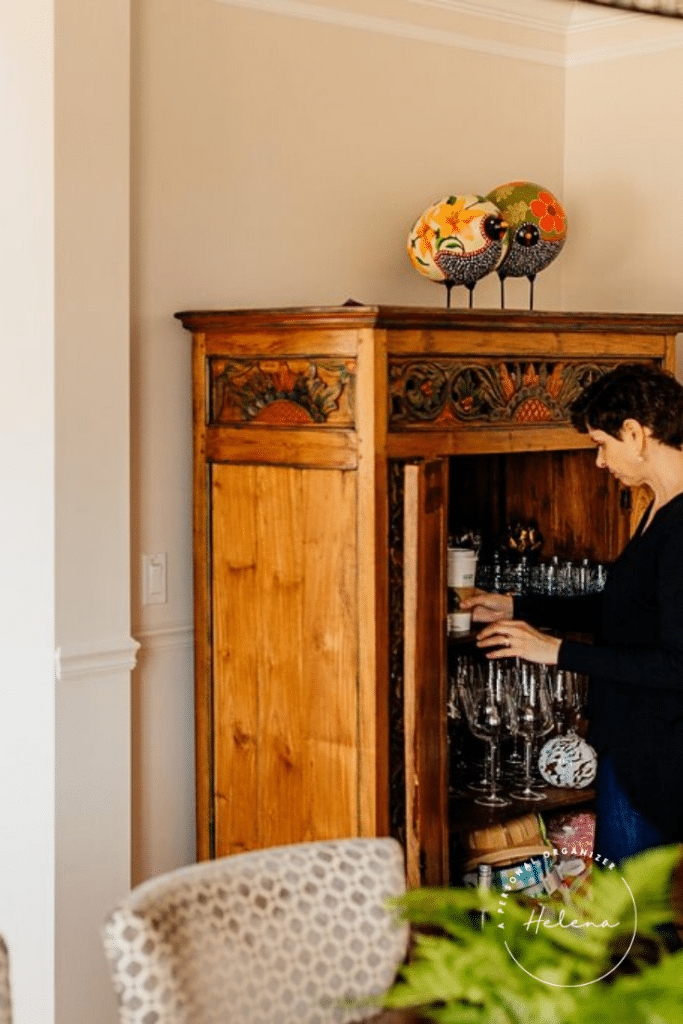 A woman in a dark sweater organizes the dining room, opening a tall, decorative wooden cabinet filled with glassware. The cabinet features painted designs and two ceramic chicken figures on top. A patterned chair is partially visible in the foreground.