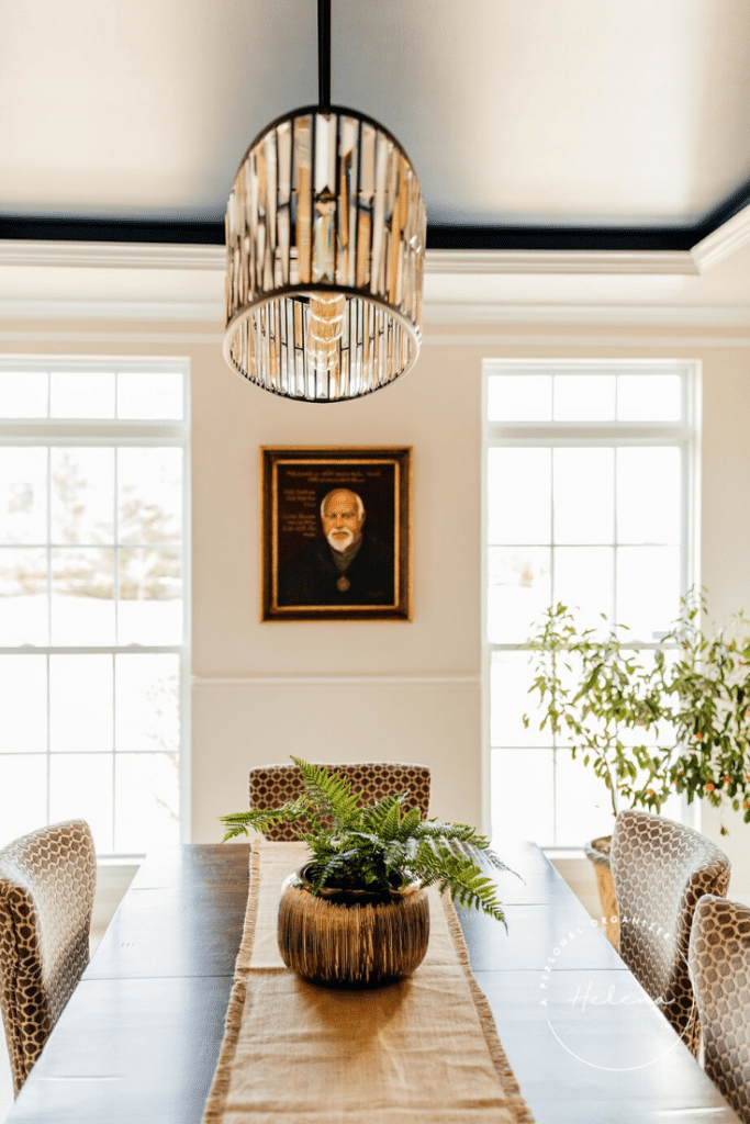Elegant dining room with a dark wood table, covered by a burlap runner and a potted fern. Meticulously organizing the dining room, leopard print chairs are neatly arranged around the table. A portrait hangs between two large windows, and a stylish overhead light fixture adds a modern touch.