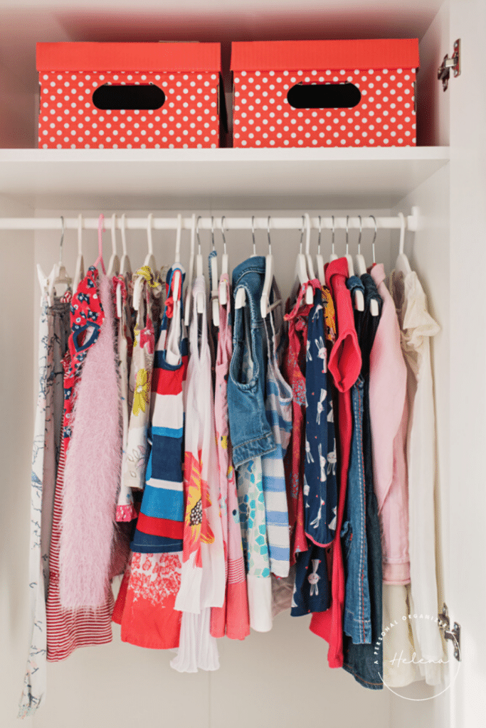 A closet beautifully organized to brighten kids' rooms, showcasing a row of colorful children's clothing on hangers with vibrant patterns in red, blue, and pink. Above the garments are two charming red polka dot storage boxes.