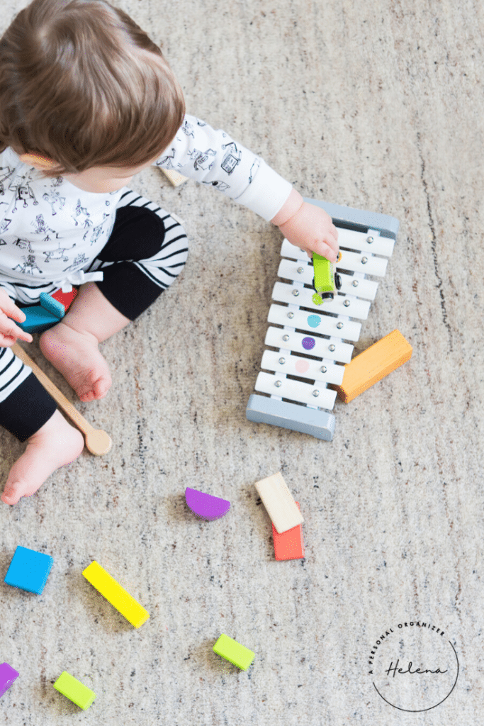 In a cozy playroom, a child sits on the carpeted floor, playing with a xylophone and colorful wooden blocks. Dressed in black and white, the child holds a green car on the xylophone while scattered blocks add to the playful chaos.