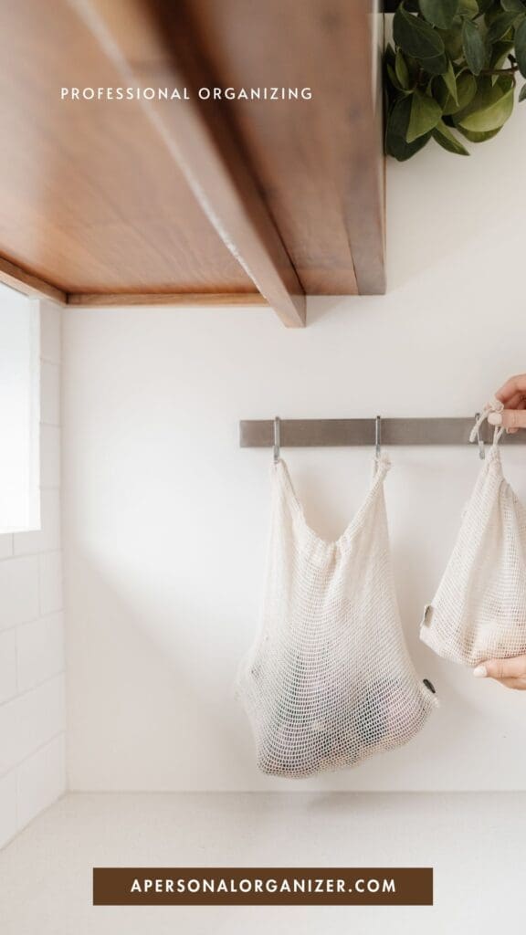 A hand hangs a reusable mesh bag on a hook under a wooden shelf in a bright, minimalist kitchen—perfect for declutter and professional organizing. Text reads "Professional Organizing" at the top and "apersonalorganizer.com" at the bottom.
