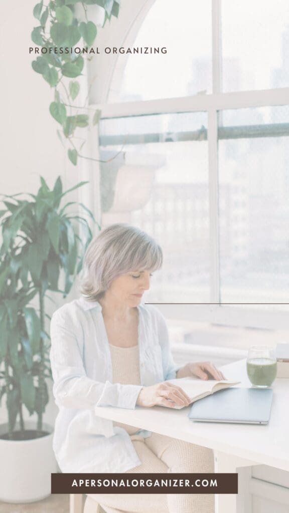 A woman with gray hair sits at a table reading a book next to a large window and green plant, enjoying organization and tidying. Text reads "Professional Organizing" and "apersonalorganizer.com." A glass of green juice is on the table.