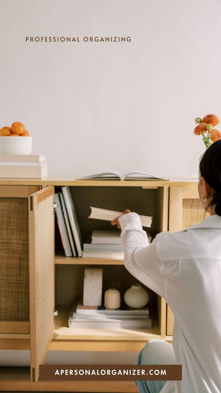 A person in a white shirt is tidying and organizing books in a light wooden cabinet with sliding doors. A bowl of oranges and small vases with flowers sit on top. Text reads "Professional Organizing" and "apersonalorganizer.com.