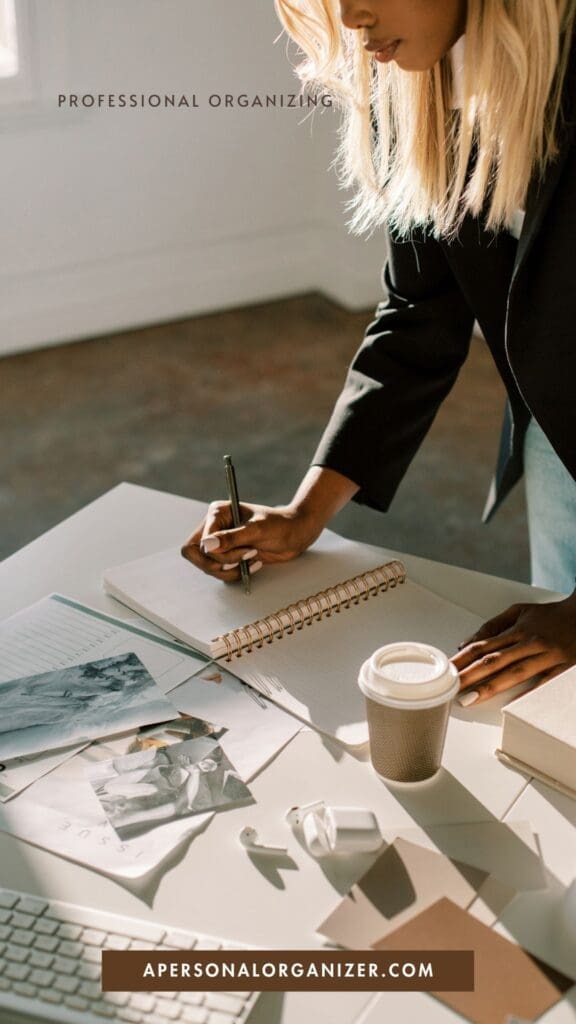A woman writes in a spiral notebook at a desk filled with clutter—papers, photos, coffee cup, glasses, and laptop—promoting expert organizing at apersonalorganizer.com.