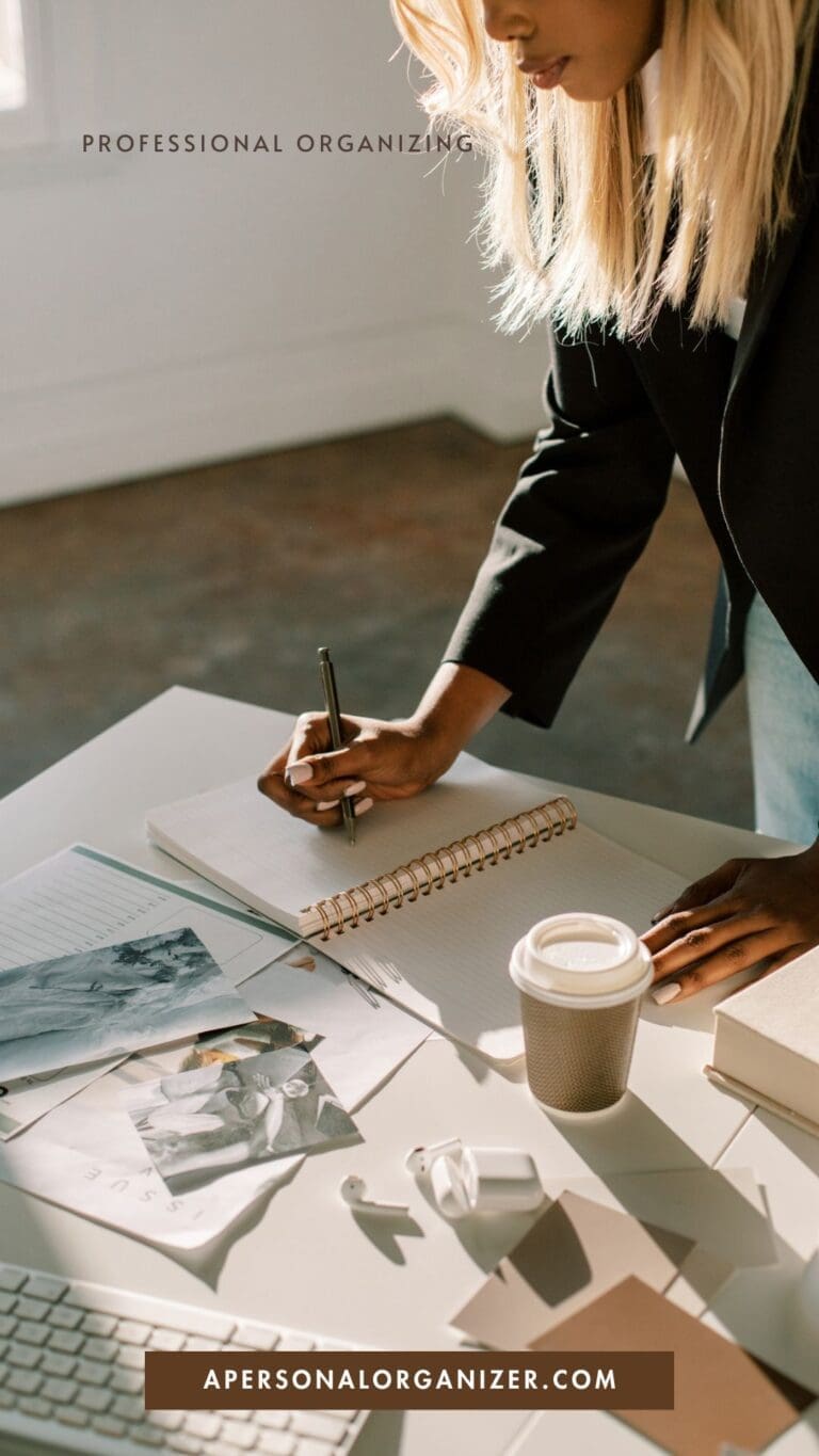 A woman writes in a spiral notebook at a desk filled with clutter—papers, photos, coffee cup, glasses, and laptop—promoting expert organizing at apersonalorganizer.com.