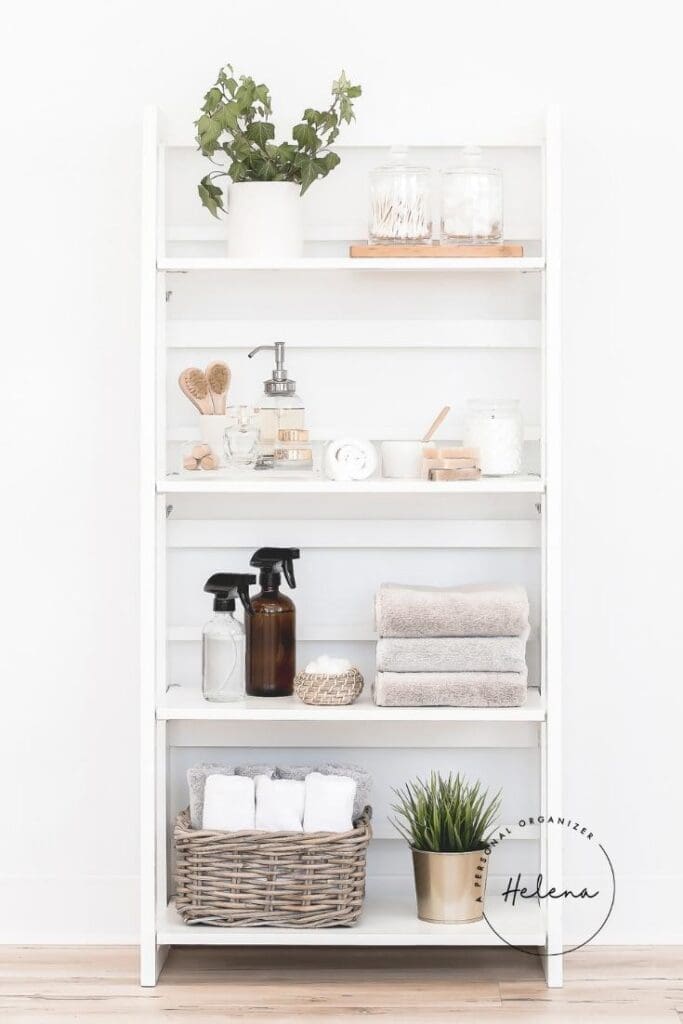 A white shelving unit serves as an organizing system, holding potted greenery, jars, brushes, soap, spray bottles, stacked towels, and a wicker basket of rolled white towels against a white wall and wooden floor.