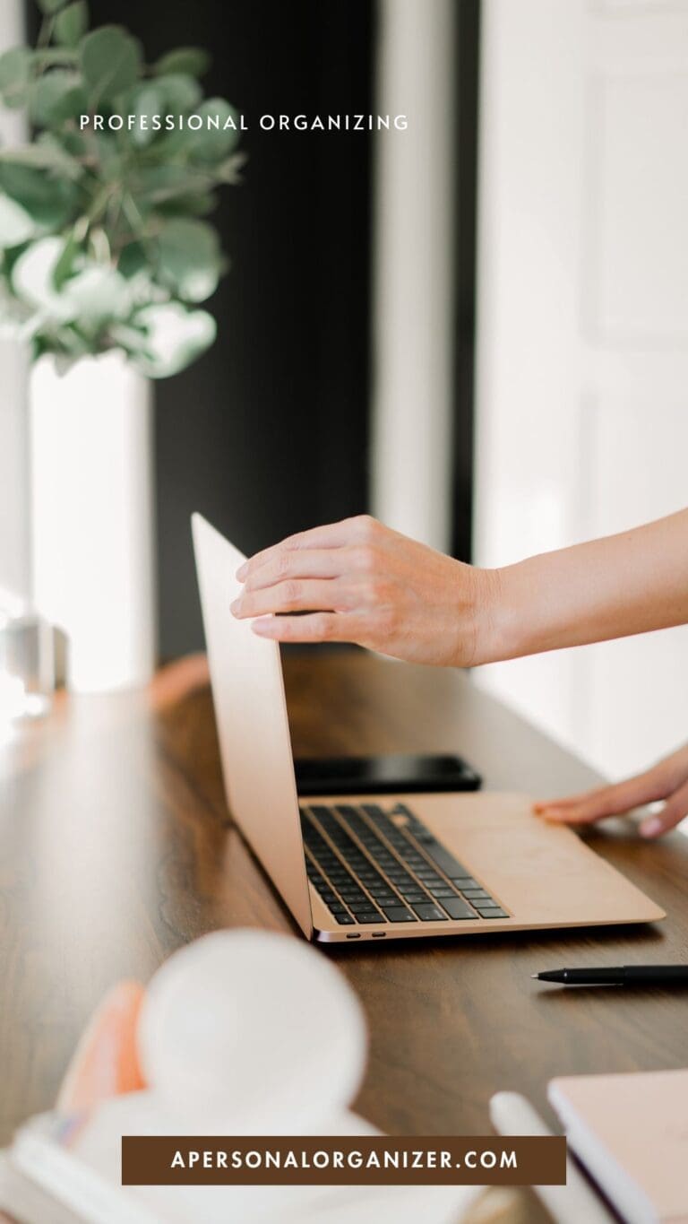 A person’s hand closes a laptop on a wooden desk with a pen, phone, and notebook nearby. Text reads "Professional Organizing" and "apersonalorganizer.com"—perfect for anyone looking to declutter and bring organization to their workspace.
