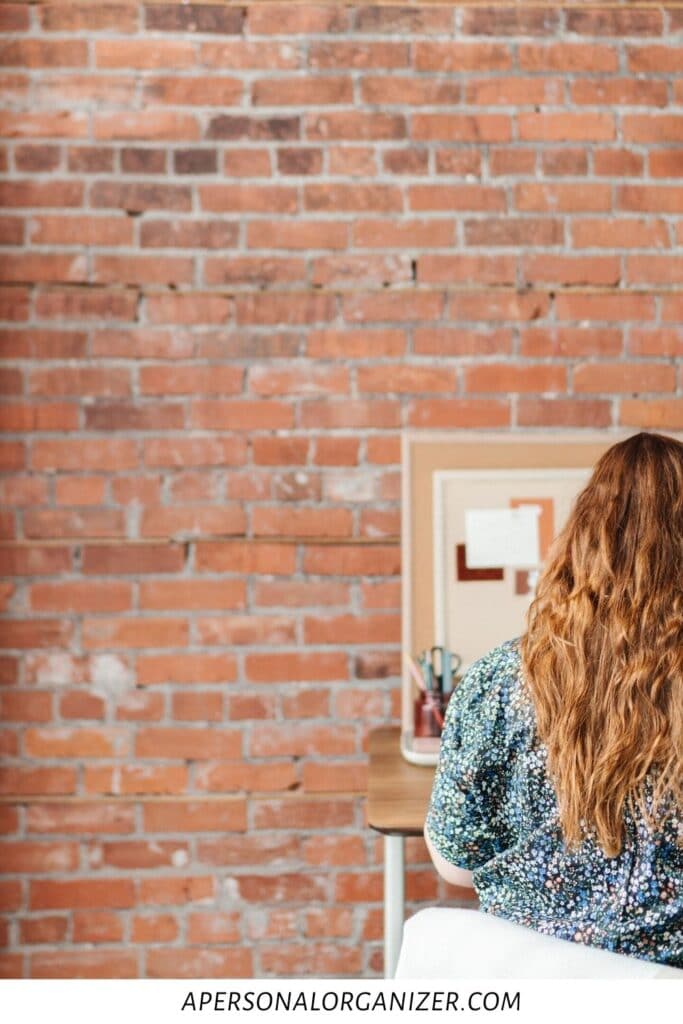 A person with long hair sits at a desk in what appears to be a casually organized college dorm room. They face a corkboard mounted on the brick wall. The website "apersonalorganizer.com" is noted at the bottom of the image, blending seamlessly into this study-friendly environment.